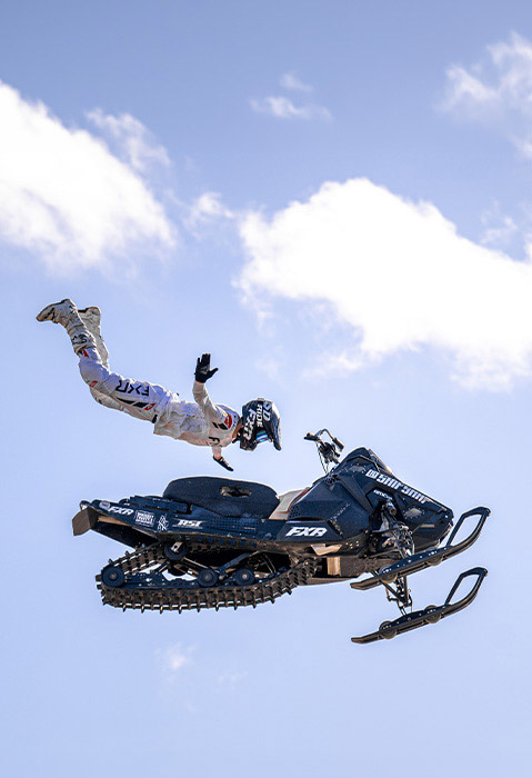 Freestyle rider performing an aerial stunt beside a snowmobile high above the ground against a bright, cloudy sky.