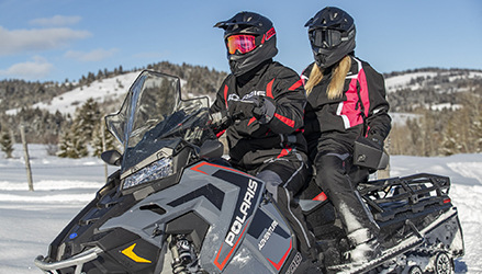 a couple riding a Polaris snowmobile sled, wearing polaris snow apparel and gear