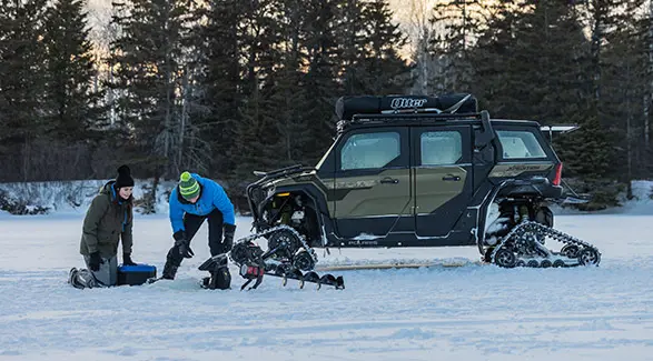2 people ice fishing with a Moss Green 2025 Polaris XPEDITION ADV 5 NorthStar side-by-side parked in the background.