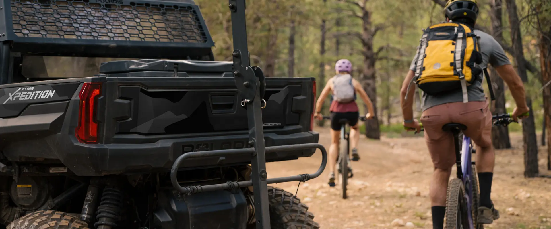 2 people on mountain bikes riding away from a 2026 Polaris XPEDITION XP NorthStar side-by-side with a bike rack installed.
