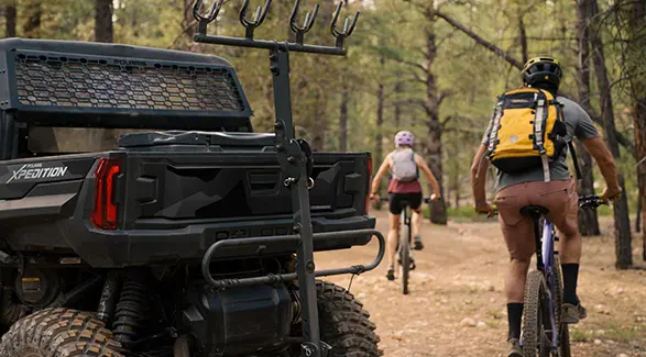 2 people on mountain bikes riding away from a 2026 Polaris XPEDITION XP NorthStar side-by-side with a bike rack installed.
