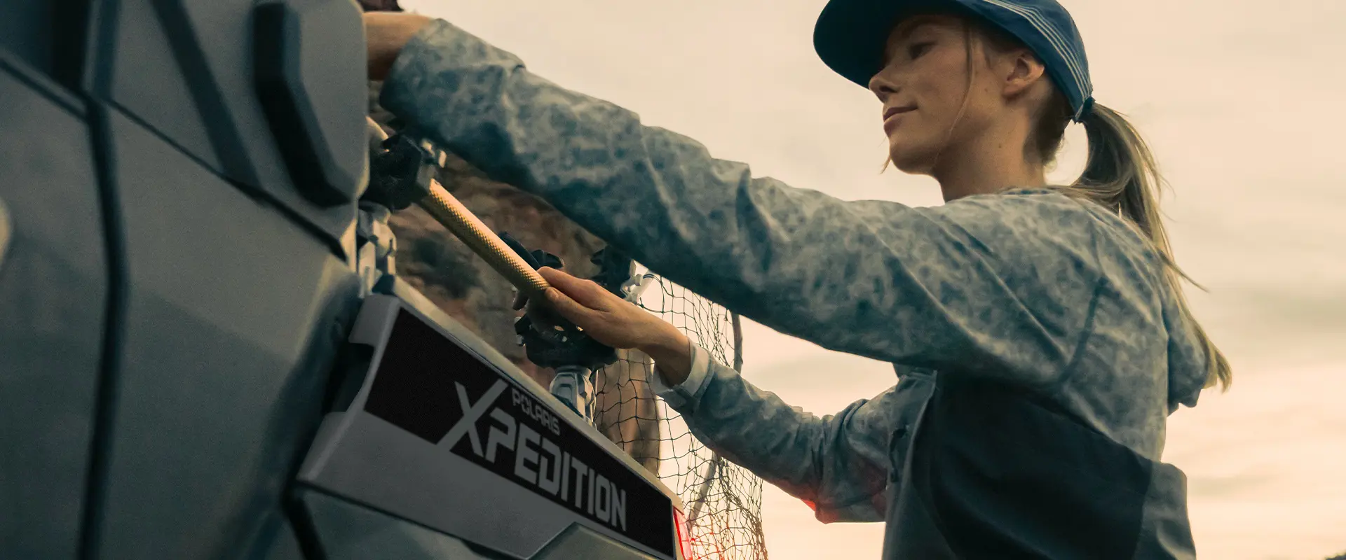 A woman putting a fishing net onto a rack installed on the cargo box of a 2026 Polaris XPEDITION XP NorthStar side-by-side.