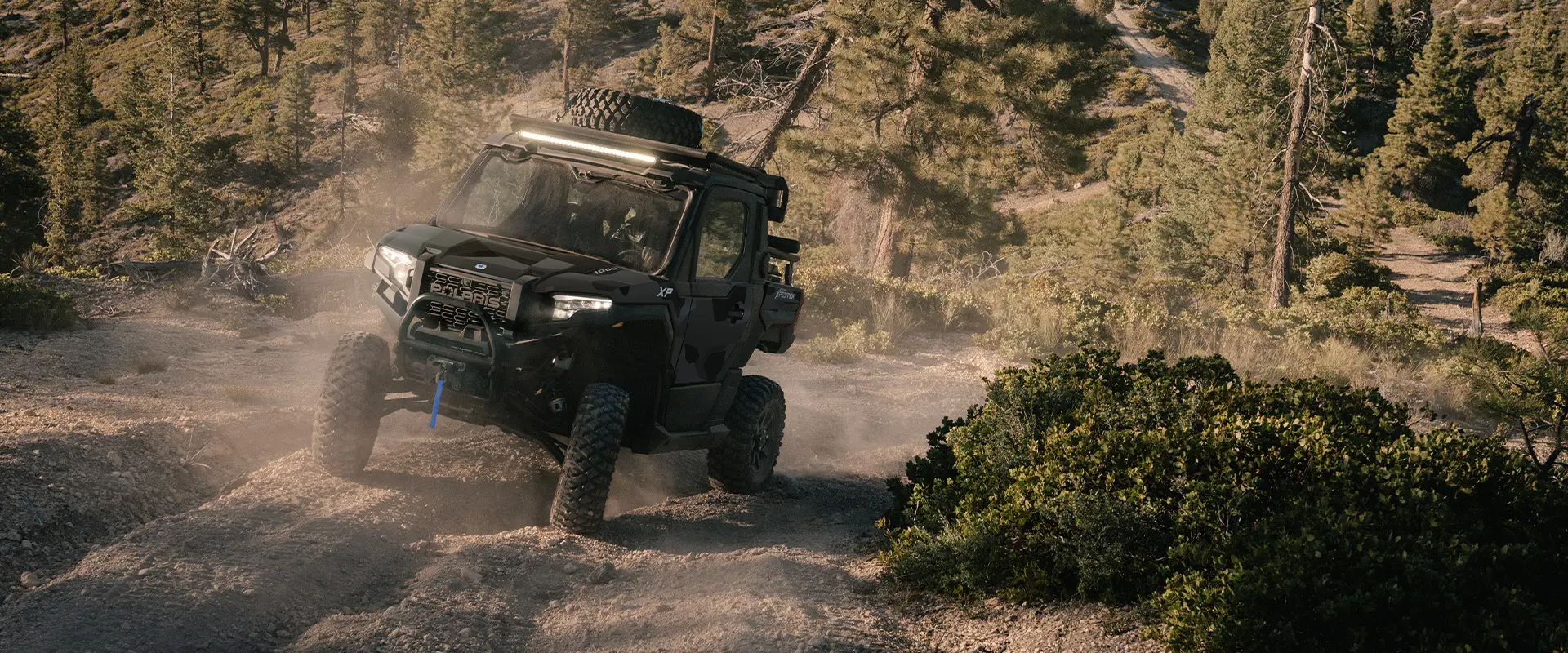 Front end view of a 2026 Polaris XPEDITION XP NorthStar side-by-side cruising on a trail with light bar and headlights illuminated.