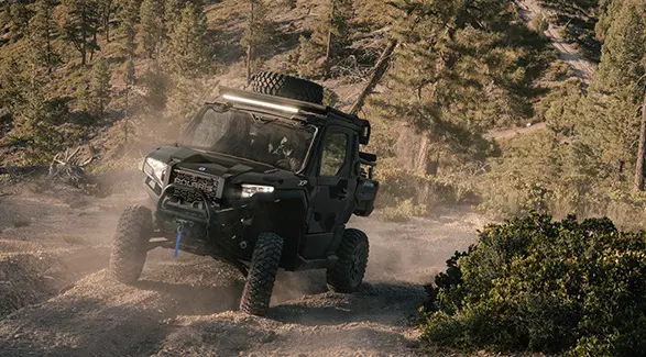 Front end view of a 2026 Polaris XPEDITION XP NorthStar side-by-side cruising on a trail with light bar and headlights illuminated.