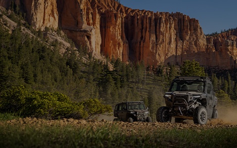 Polaris XPEDITION XP Side-by-Side leading a Polaris XPEDITION ADV 5 SxS along a rocky mountain trail, kicking up dust with pine forests and canyon walls in the background.